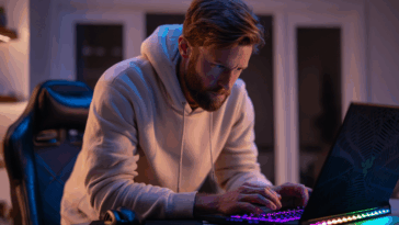 Jeune homme de 25 ans en train de brancher son ordinateur portable gaming sur un bureau, concentré, clavier rétroéclairé, chaise gamer en arrière-plan.